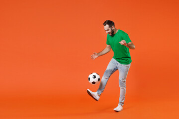 Full length portrait funny young man football fan in basic green t-shirt cheer up support favorite team kicking juggling soccer ball isolated on orange background studio. People sport leisure concept. - obrazy, fototapety, plakaty