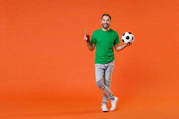 Fototapeta na wymiar Full length portrait funny young man football fan in green t-shirt cheer up support favorite team with soccer ball hold beer bottle isolated on orange background studio. People sport leisure concept.