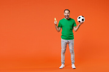 Fototapeta na wymiar Full length portrait of cheerful man football fan in basic green t-shirt cheer up support favorite team with soccer ball using mobile phone isolated on orange background. People sport leisure concept.