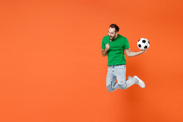 Fototapeta na wymiar Full length portrait crazy man football fan tearing green t-shirt cheer up support favorite team with soccer ball jumping screaming isolated on orange background studio. People sport leisure concept.