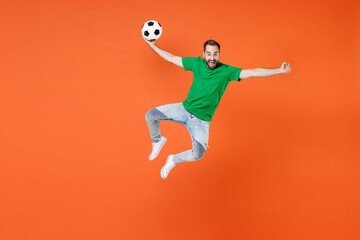 Fototapeta na wymiar Full length portrait of excited man football fan in green t-shirt cheer up support favorite team with soccer ball jumping clenching fist isolated on orange background. People sport leisure concept.