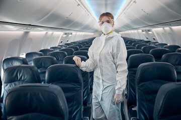 Female flight attendant standing on the salon of civil airplane in protective glasses