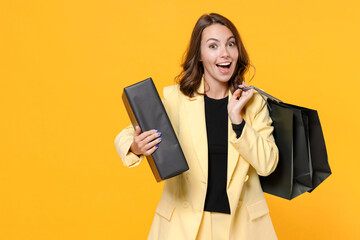 Surprised young brunette woman 20s wearing basic light suit jacket hold package bags box with purchases after shopping looking camera isolated on yellow background, studio portrait. Black friday sale.