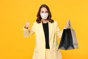 Fototapeta na wymiar Young woman in suit jacket face mask to safe from coronavirus virus covid-19 hold package bags with purchases after shopping showing thumb up isolated on yellow background studio. Black friday sale.
