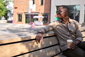 positive black male drink coffee outdoors, take a break after hard working day. sit on bench