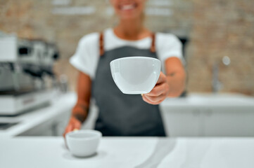 Young attractive female barista stands at the counter in a coffee shop and smiles, serving cups of prepared coffee to a customer.