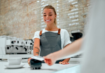 Young attractive female barista stands at the counter in a coffee shop. Customer paying for their order with a credit card in a cafe.
