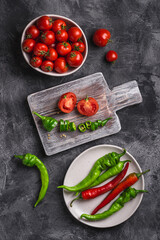 Fresh ripe tomatoes in bowl near to old wooden cutting board with hot chili peppers in plate, dark stone concrete background, top view