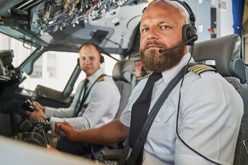 Serious bearded pilot sitting in the cockpit of passenger aircraft - obrazy, fototapety, plakaty