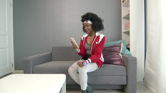 Young african american woman hippy in colourful sportswear with curly afro hairs lifts dumbbell and uses phone to make stories or photo