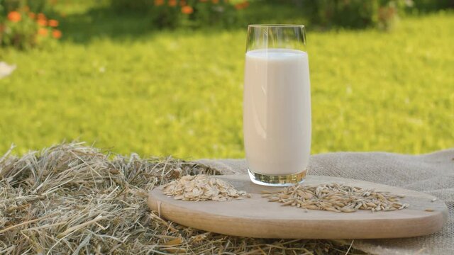 Close up oat milk in a glass. Oat grains and oatmeal on a wooden cutting board. Fresh hay, jute burlap, rural still life. The camera moves slowly on the slider