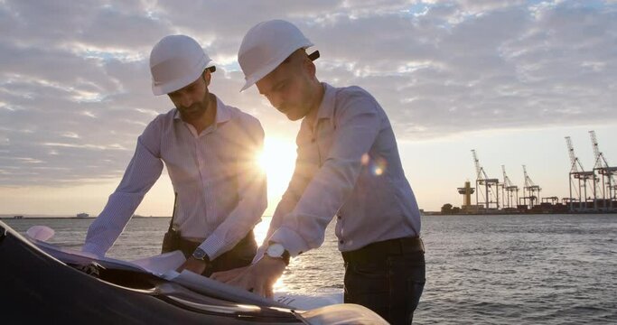 Engineers in white helmets work with paper drawings laid out on the hood of a car in front of the seaport at dawn.