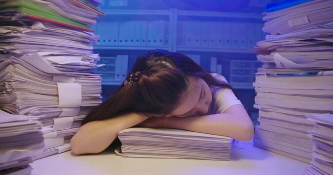 Tired young asian student woman lay down on desk cover with stack of paperwork. Alone teen girl sleep on pile of paper sheet while studying hard at late night. Smoke on background. Dolly shot.