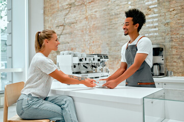 Attractive African male professional barista in apron serves a cup of coffee to a client and smiles.