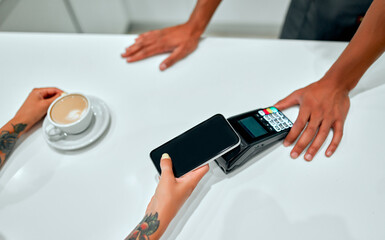 Obraz premium Female customer making payment through mobile phone at counter in cafe with young man. Barista holding credit card reading machine in front of female costumer with cell phone.