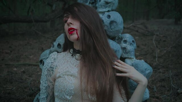 portrait of a young woman vampire, beautiful red lips with dripping blood drops on her face, sharp false fangs. A model in a dark gothic forest against the backdrop of the scenery of human skulls.