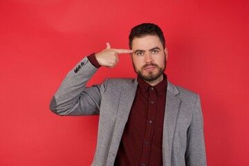 Unhappy Young caucasian businessman wearing casual clothes standing  makes suicide gesture and imitates gun with hand, curves lips keeps two fingers on temple, shoots, being tired of everything,