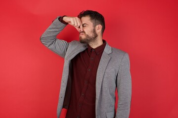 Young caucasian businessman wearing casual clothes standing over isolated red background smelling something stinky and disgusting, intolerable smell, holding breath with fingers on nose. Bad smell