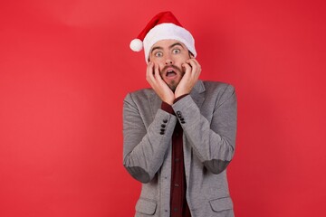 Stupefied Young caucasian businessman wearing casual clothes standing over isolated red background expresses excitement and thrill, keeps jaw dropped, hands on cheeks, has eyes popped out