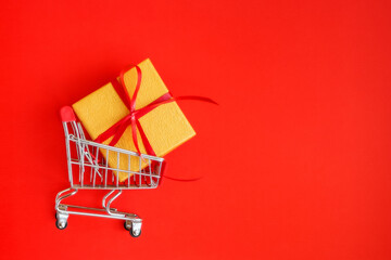 Shopping cart, small gift box on a red background with a copy of the space. Christmas online shopping.