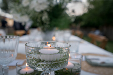 Beautiful decoration of a wedding banquet in the green garden in white colors. Serving the table with white tablecloth, candle, plates, glasses, cutlery, napkins decoration, with white flowers in vase