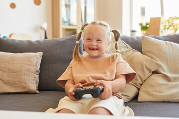Portrait of cute girl with down syndrome playing video games and laughing happily while sitting on...