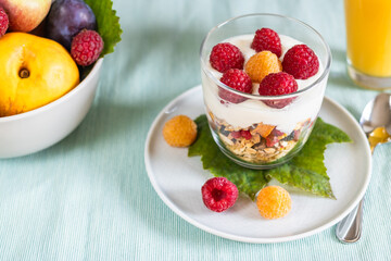 Close up of a dessert with granola, natural yoghurt and raspberries and fruit and orange juice in the background