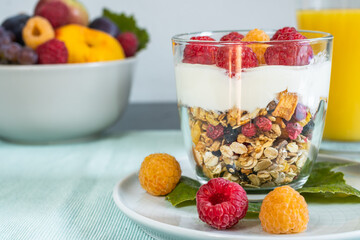 Close up of a dessert with granola, natural yoghurt and raspberries and a bowl full of fruit and orange juice in the background