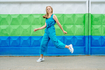 beautiful happy girl in blue long jumpsuit having fun near the orange wall. The girl jumps and runs in a hurry