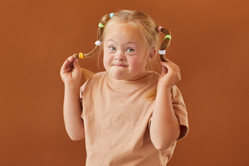 Waist up portrait of cute blonde girl with down syndrome making faces at camera while standing...