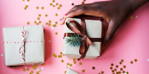 African woman's hand taking a craft textured gift box with ribbon bow surrounded by stars and snowflakes on a coral background, from above