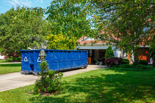 An empty blue dumpster in the driveway of a house with its garage door open in a residential community