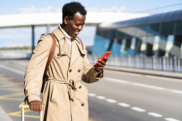 Smiling Afro-American traveler man with suitcase stands in airport terminal, using mobile application in smart phone to rent vehicle online in car sharing service, calling taxi. Rental concept. 