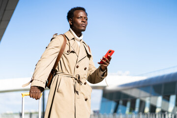 Afro-American millennial traveler man with yellow suitcase stands in airport terminal, holding phone, calling and looking for a taxi or car sharing. Black male waiting for cab for transporting
