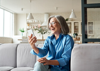 Happy mature old 60s woman holding smartphone using mobile phone app for video call, laughing while...