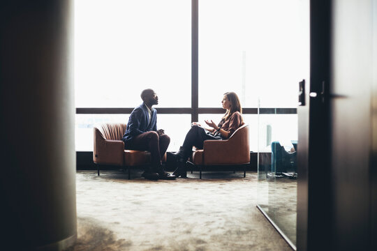 Male and female colleagues discussing while sitting against window at workplace