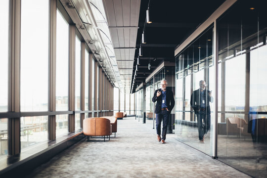 Mature businessman using smart phone while walking in corridor at workplace