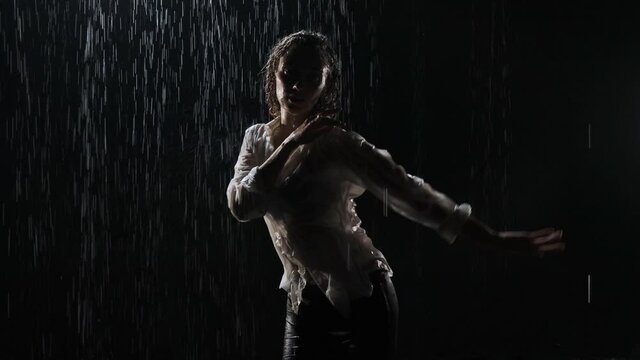 Beautiful young woman dancing in a white shirt and black short shorts dancing in the rain in the studio on a black background. Dancing on the water.