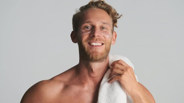 Attractive smiling blond bearded man with eye patches and towel on shoulder confidently looking in camera over white background