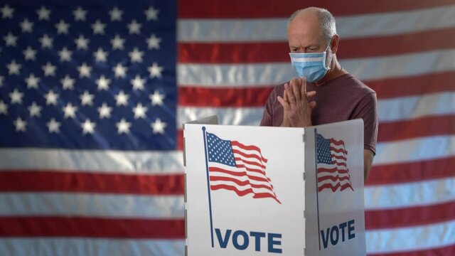 Conceptual portrait on US flag background of man in voting booth at ballot box wearing face mask, careful decision for president, with thumbs up for US election during pandemic.