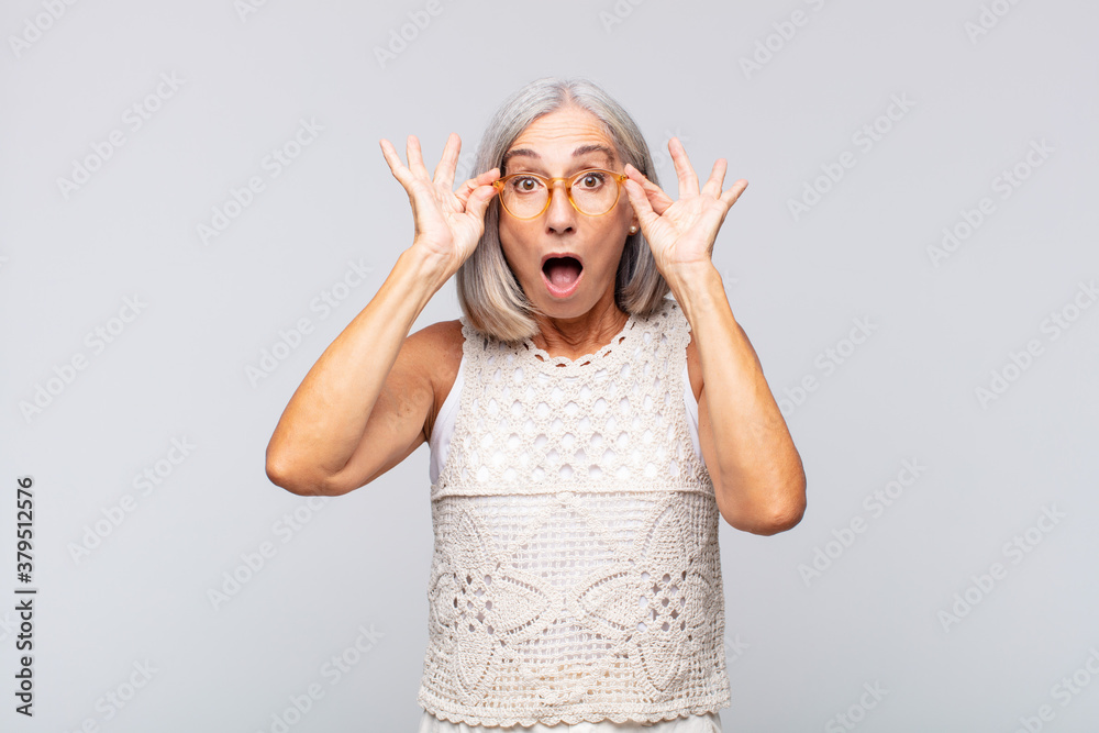 Wall mural gray haired woman feeling shocked, amazed and surprised, holding glasses with astonished, disbelieving look