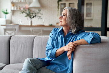 Calm relaxed mature older woman relaxing sitting on couch at home. Peaceful middle aged grey-haired lady resting on sofa in modern living room enjoying lounge and no stress, looking away, thinking. - obrazy, fototapety, plakaty