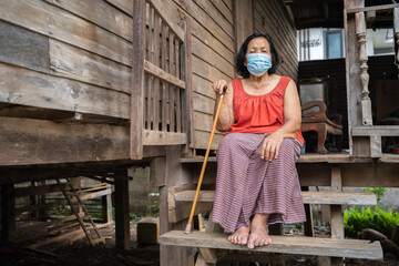 Thai elderly woman in round-necked sleeveless collar wearing medical mask for protect corona virus (covid-19) pandemic in wooden home