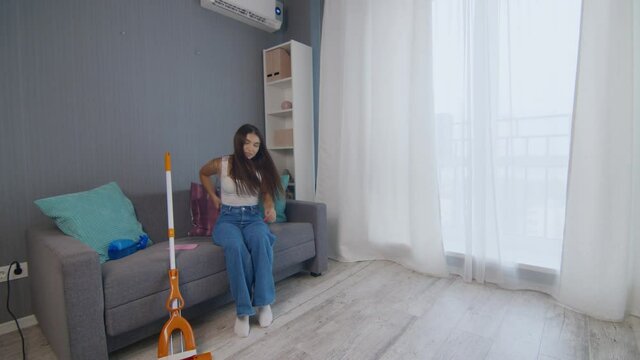 Tired young woman after cleaning the house alone sits on coach and uses phone