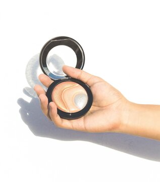 Manos de mujer sosteniendo maquillaje iluminador, luz dura fondo blanco con sombra