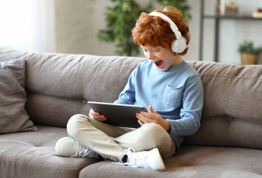 Delighted redhead boy with tablet sitting on sofa.