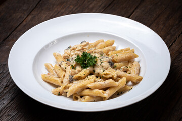 Homemade delicious pasta macaroni with white mushrooms chicken meet mushrooms in creamy cheese sauce in the white plate on the dark brown wood table. Parsley, spicy. Yellow, green.