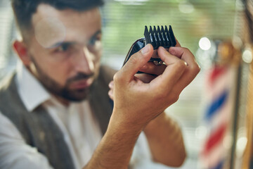Professional barber looking attentively at an electric hair trimmer - obrazy, fototapety, plakaty