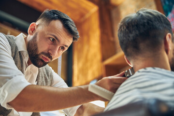 Focused young barber using electric hair trimmer in his work