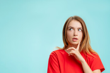 Curious woman. Special offer. Portrait of doubtful lady in red t-shirt with finger on chin thinking with open mouth looking up at copy space isolated on blue promotional background. Inspiration idea.